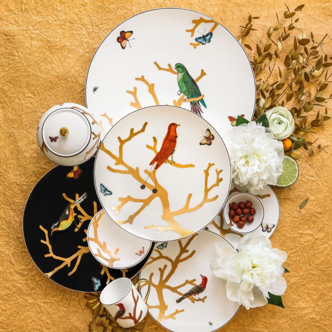 The Aux Oiseaux Dinnerware by Bernardaud showcases plates, cups, and a teapot adorned with vibrant birds and butterflies on branches, displayed on gold with white peonies, greenery, and berries.