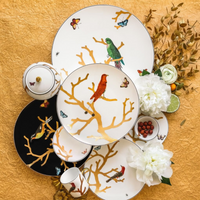 The Aux Oiseaux Dinnerware by Bernardaud showcases plates, cups, and a teapot adorned with vibrant birds and butterflies on branches, displayed on gold with white peonies, greenery, and berries.