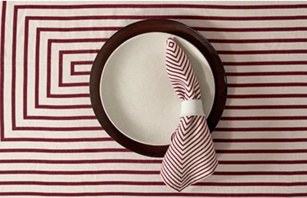 A round Terra Stone Dinnerware plate by L'Objet with a dark rim sits on a geometric red-striped tablecloth, topped with a folded red-and-white striped napkin in a napkin ring.