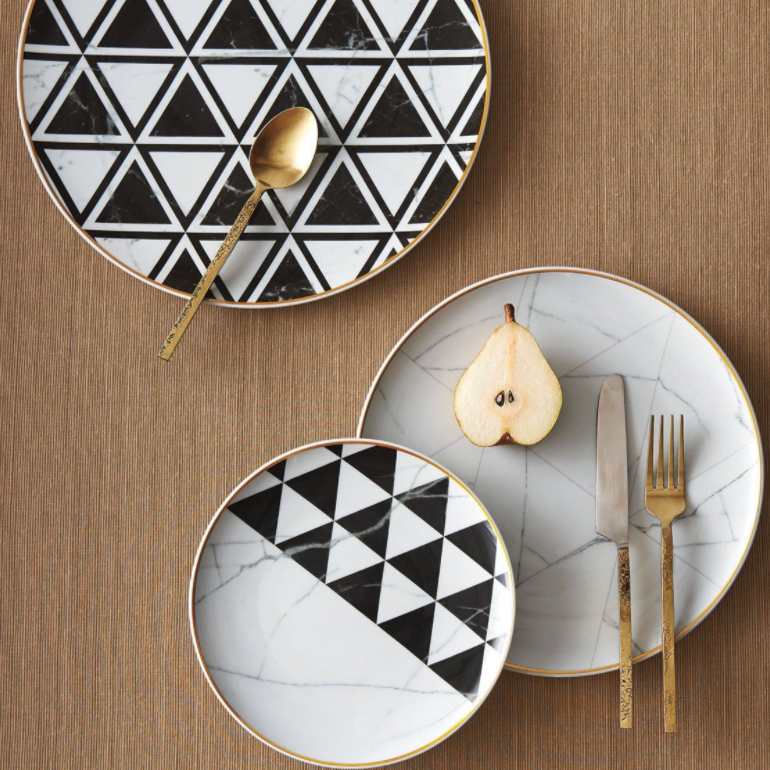 Three dinnerware plates from Carrara by Coline Le Corréa for Vista Alegre rest on a woven placemat: two with black and white geometric triangles, one with a marble pattern. A gold fork, knife, spoon, and halved pear complete this designer-inspired setting.