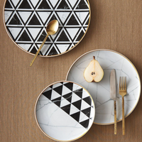 Three dinnerware plates from Carrara by Coline Le Corréa for Vista Alegre rest on a woven placemat: two with black and white geometric triangles, one with a marble pattern. A gold fork, knife, spoon, and halved pear complete this designer-inspired setting.