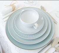 A place setting features a large blue Venezia Dinnerware plate by Vista Alegre, two patterned white porcelain plates, and a matching teacup stacked on top. Silverware surrounds the plates, and a rolled napkin is tied with string.