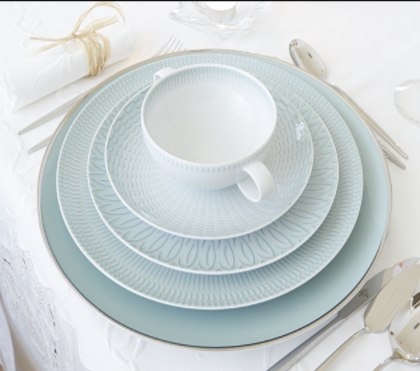 A place setting features a large blue Venezia Dinnerware plate by Vista Alegre, two patterned white porcelain plates, and a matching teacup stacked on top. Silverware surrounds the plates, and a rolled napkin is tied with string.