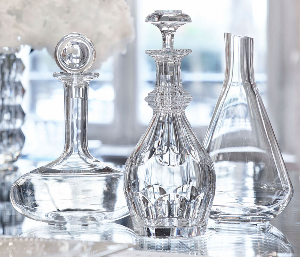 Three Baccarat Chateau Decanters are elegantly displayed on a reflective surface, illuminated by soft natural light. Each decanter showcases a distinct shape and intricate design, highlighting Baccarat’s renowned craftsmanship.