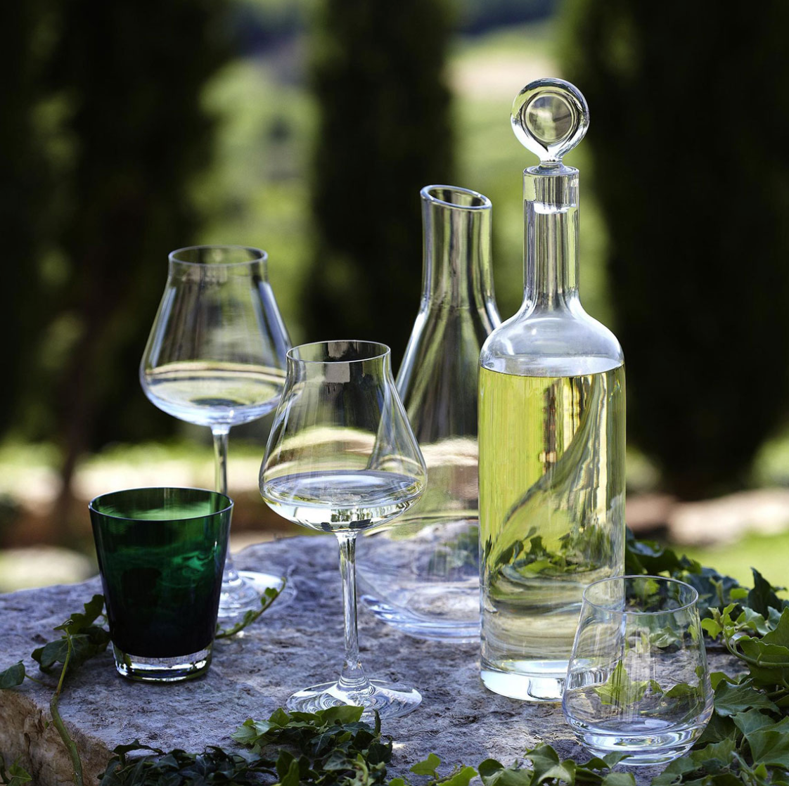 A Baccarat Chateau Decanter with matching clear wine glasses, a tumbler, and a green glass sit on a stone surface outdoors, surrounded by lush greenery and tall trees in the background.