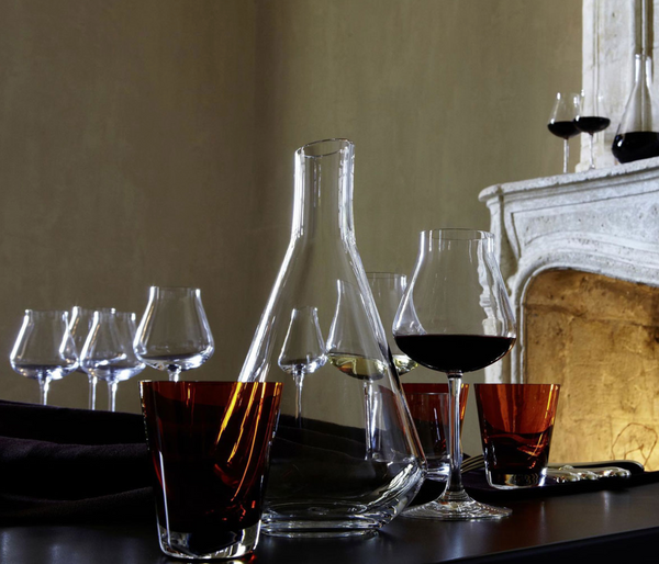 A table set with empty wine glasses, a Baccarat Chateau Decanter, and several filled glasses—some with red wine, others amber. In the background, more wine glasses decorate a fireplace.