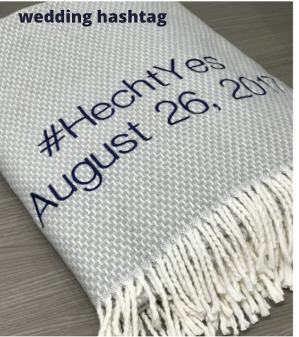 A Soft Idea’s Personalized Throw Blanket: folded white with fringe, embroidered in dark blue with “#HechtYes August 26, 2017.” Above the blanket, “wedding hashtag” appears in black text.