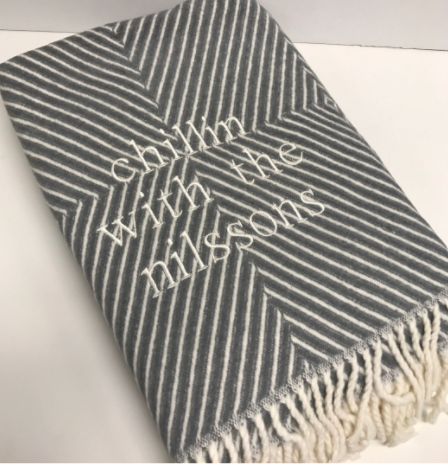 A Soft Idea’s Personalized Throw Blanket features a folded gray and white chevron pattern, white fringe, and “chillin with the nilssons” embroidered in white thread.