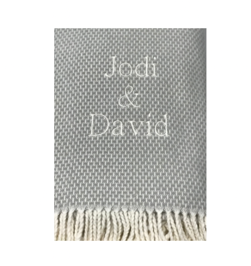 A close-up of the A Soft Idea Personalized Throw Blanket in gray, featuring white fringe and “Jodi & David” embroidered in white thread at the center.
