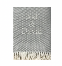 A close-up of the A Soft Idea Personalized Throw Blanket in gray, featuring white fringe and “Jodi & David” embroidered in white thread at the center.