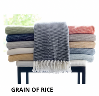A stack of colorful woven blankets rests on a white cushioned bench. A dark gray A Soft Idea Personalized Throw Blanket with white fringes drapes over the front. The words GRAIN OF RICE appear beneath it.
