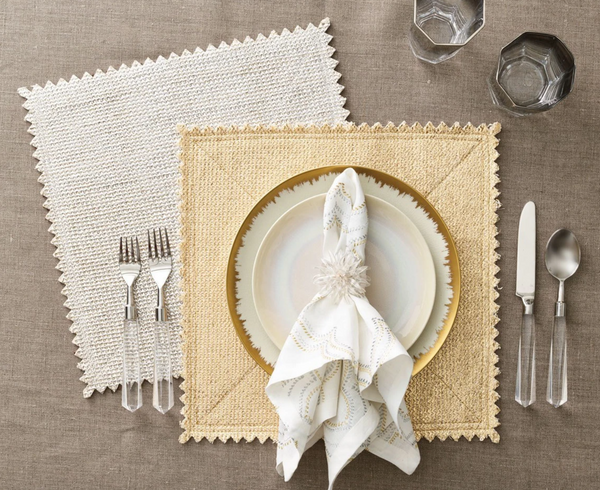 Two square Stamped Reversible Placemat Silver & Gold placemats with scalloped edges arranged on a table with a place setting of plates, a napkin in a ring, flatware, and two glasses.