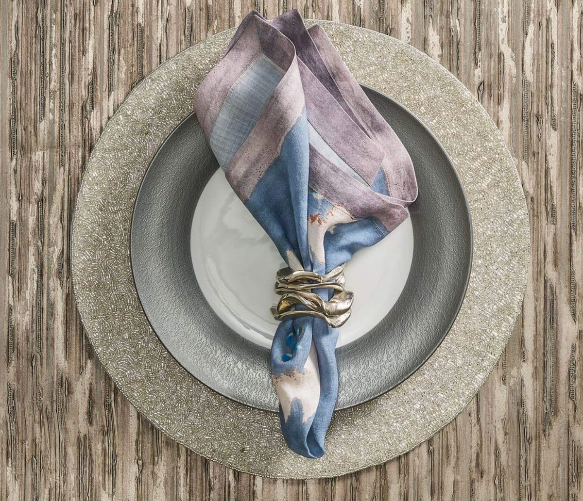 A white plate on a silver charger atop a wooden table is accented by a blue and purple cloth napkin with a Kim Seybert Flux Napkin Ring Silver, adding an elegant touch to the table setting.
