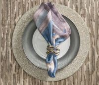 A white plate on a silver charger atop a wooden table is accented by a blue and purple cloth napkin with a Kim Seybert Flux Napkin Ring Silver, adding an elegant touch to the table setting.