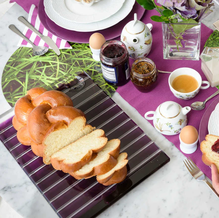A breakfast table set on a purple cloth showcases braided bread on the Apeloig Striped Acrylic Challah Board & Knife, alongside jam jars, tea, boiled eggs in holders, floral napkins, a vase of purple flowers, and neatly arranged plates and cutlery.