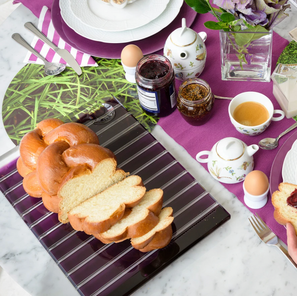 A breakfast table set on a purple cloth showcases braided bread on the Apeloig Striped Acrylic Challah Board & Knife, alongside jam jars, tea, boiled eggs in holders, floral napkins, a vase of purple flowers, and neatly arranged plates and cutlery.