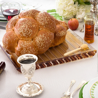 A round challah sits on the Apeloig Striped Acrylic Challah Board & Knife, joined by a silver wine goblet, honey jar with dipper, apple, and elegant tableware—setting the stage for a festive meal.