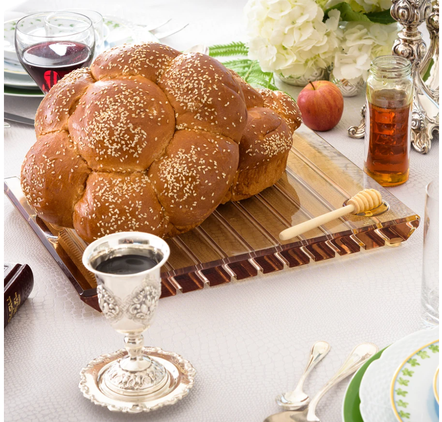 A round challah with sesame seeds rests on the Apeloig Striped Acrylic Challah Board & Knife | Various, surrounded by a honey jar, dipper, silver wine cup, plates, utensils, and a peach in an elegant table setting.