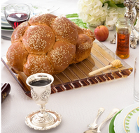 A round challah with sesame seeds rests on the Apeloig Striped Acrylic Challah Board & Knife | Various, surrounded by a honey jar, dipper, silver wine cup, plates, utensils, and a peach in an elegant table setting.