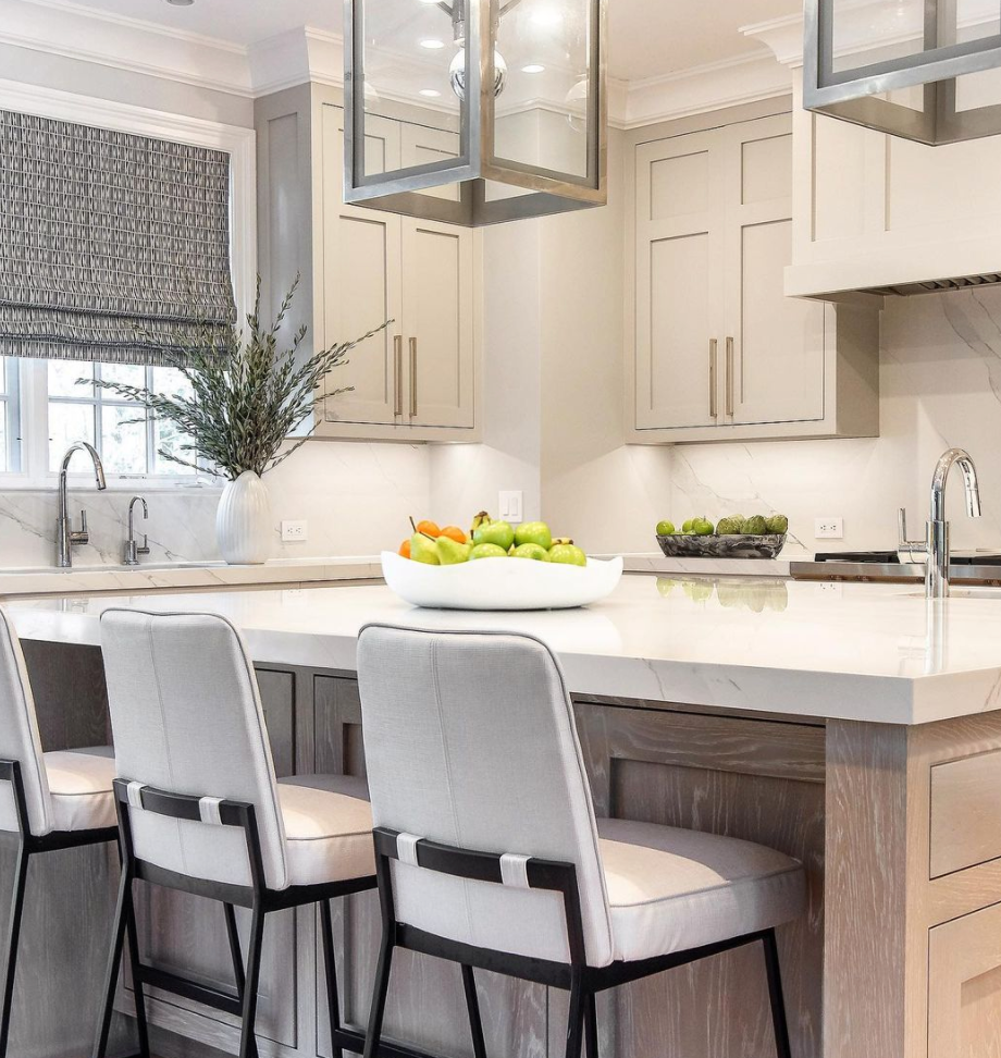 A modern kitchen with light cabinetry, a marble island with three cushioned chairs, and a Global Views/Surya Organic Round Bowl Matte White holding apples and oranges. A vase with greenery sits near a window with patterned blinds.