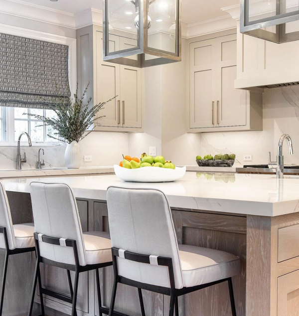 A modern kitchen with light cabinetry, a marble island with three cushioned chairs, and a Global Views/Surya Organic Round Bowl Matte White holding apples and oranges. A vase with greenery sits near a window with patterned blinds.