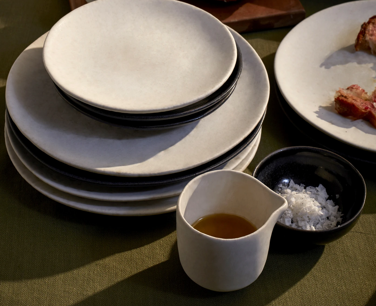 A stack of L'Objet Terra Stone Dinnerware plates sits beside a small bowl of flaked salt and a sauce jug on a green tablecloth, with a partially visible plate of food in the background.