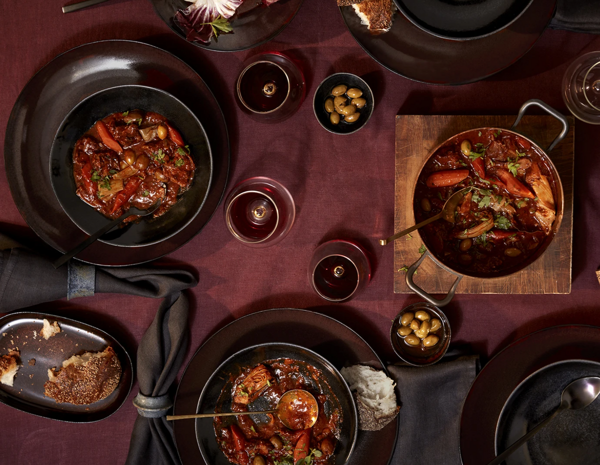 A table set with L'Objet Terra Wine Dinnerware, black plates, bowls of beef stew, carrots, olives, rustic bread, and red wine on a burgundy tablecloth. Cloth napkins and handcrafted details create an elegant setting.