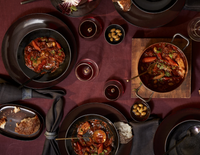 A table set with L'Objet Terra Wine Dinnerware, black plates, bowls of beef stew, carrots, olives, rustic bread, and red wine on a burgundy tablecloth. Cloth napkins and handcrafted details create an elegant setting.