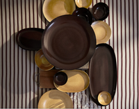 The Terra Wine Dinnerware by L'Objet, with handcrafted round and oval pieces in brown and gold, is arranged on a striped tablecloth alongside two clear glasses and a partially visible wooden board.