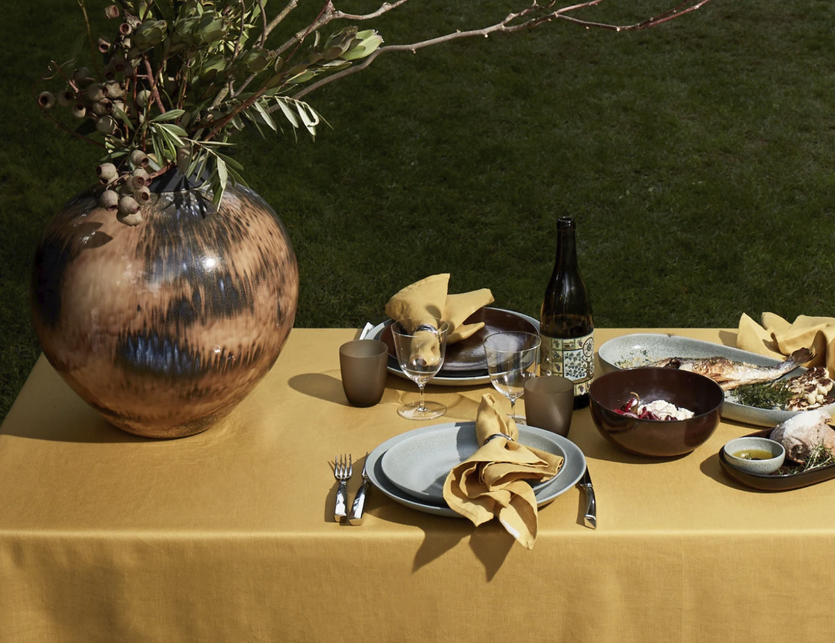 Outdoors on grass, a table dressed in yellow features L'Objet’s Terra Wine Dinnerware, cutlery, glasses, a wine bottle, napkins, and a large ceramic vase with branches. Plates are set with food for an inviting al fresco meal.