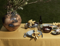 Outdoors on grass, a table dressed in yellow features L'Objet’s Terra Wine Dinnerware, cutlery, glasses, a wine bottle, napkins, and a large ceramic vase with branches. Plates are set with food for an inviting al fresco meal.