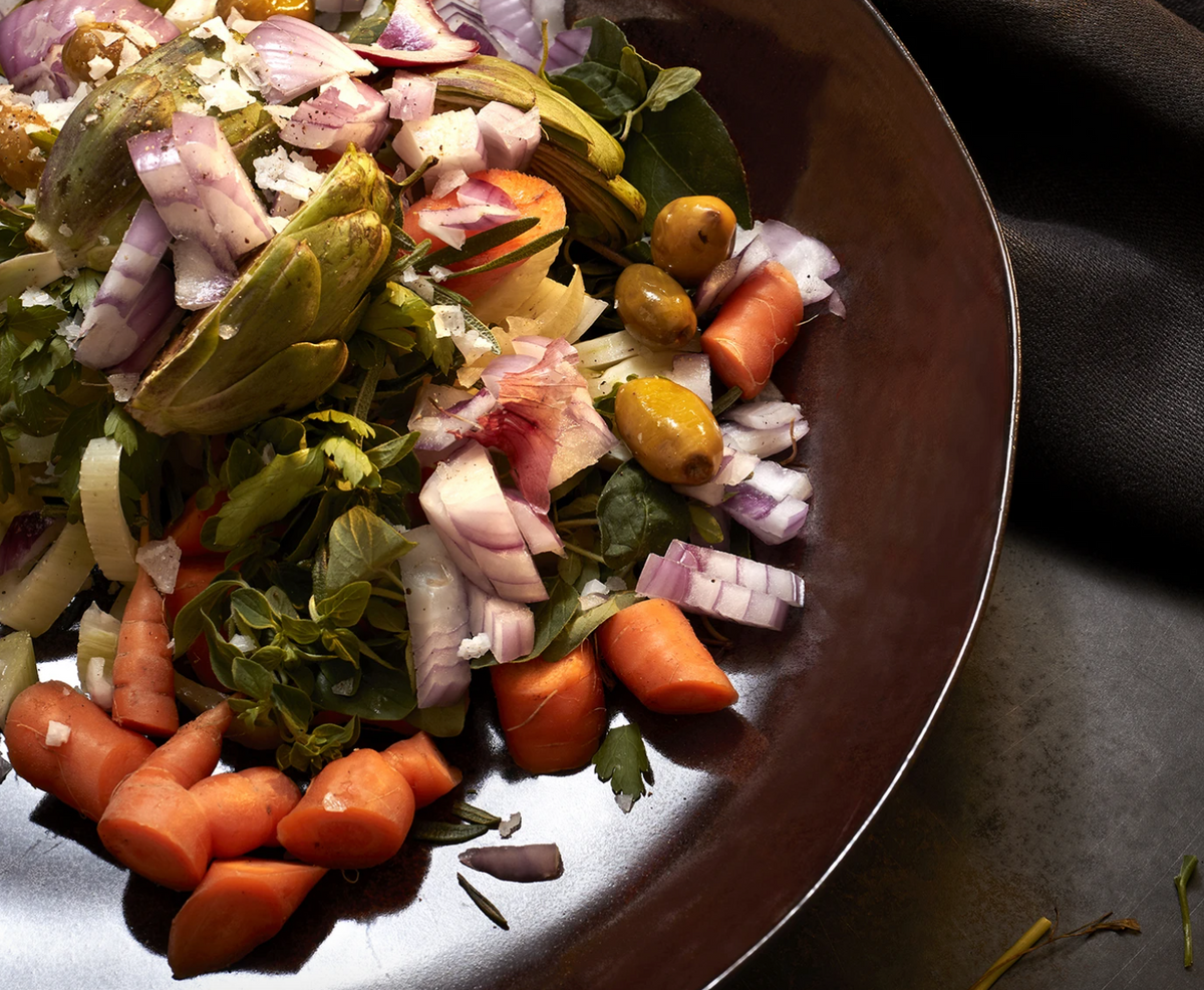 A close-up of a colorful salad with carrots, red onions, olives, artichokes, and leafy greens served on the L'Objet Terra Wine Dinnerware, with a dark cloth napkin nearby.