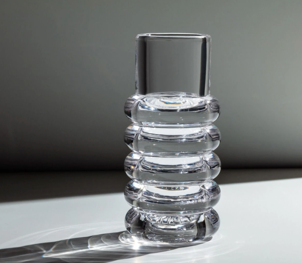 The Tom Dixon Press Stem Vase, by Tom Dixon, features clear cylindrical glass with four rounded ridges and sits on a flat surface, casting a soft shadow in diffused light for a modern look.