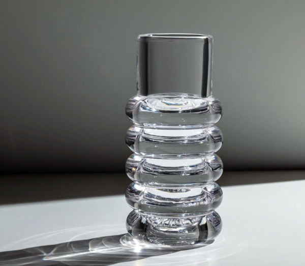 The Tom Dixon Press Stem Vase, by Tom Dixon, features clear cylindrical glass with four rounded ridges and sits on a flat surface, casting a soft shadow in diffused light for a modern look.