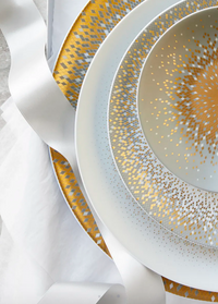 A close-up of stacked Souffle D'or Dinnerware by Haviland, featuring gold and silver geometric patterns on porcelain, displayed on a yellow and white charger with a white satin ribbon.