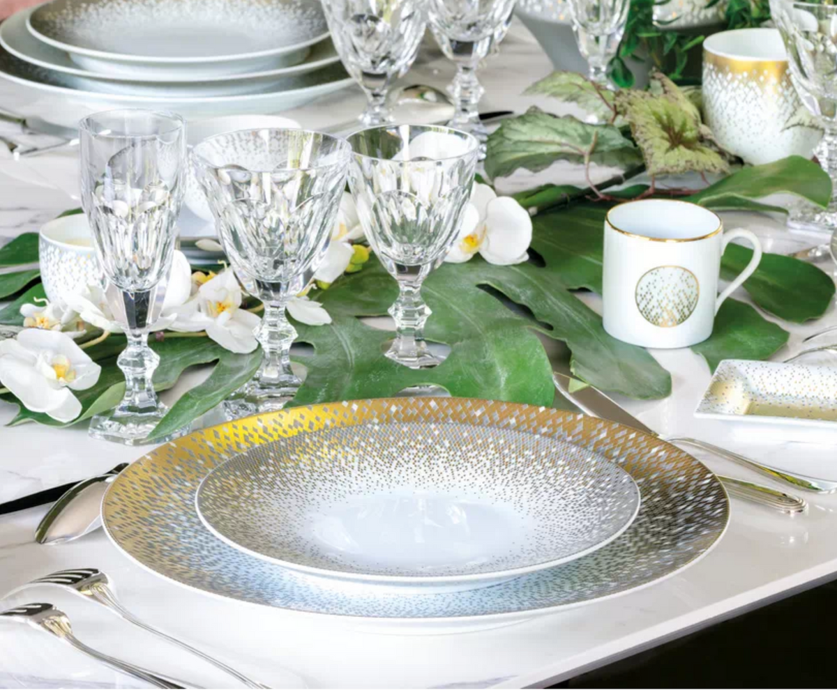 A sophisticated table setting featuring Haviland's Souffle D'or Dinnerware with gold and silver accents, crystal glasses, lush green leaves, and white flowers for a festive atmosphere.