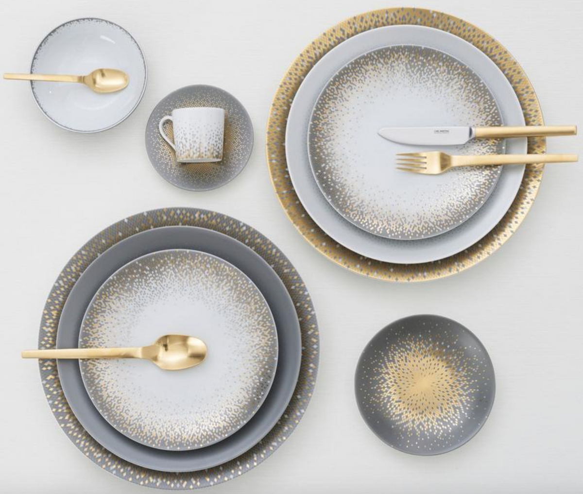 The Haviland Souffle D'or Dinnerware set includes handmade grey and white plates, bowls, a cup with gold speckled patterns, and matching gold cutlery, all elegantly arranged on a white surface.