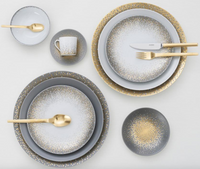 The Haviland Souffle D'or Dinnerware set includes handmade grey and white plates, bowls, a cup with gold speckled patterns, and matching gold cutlery, all elegantly arranged on a white surface.