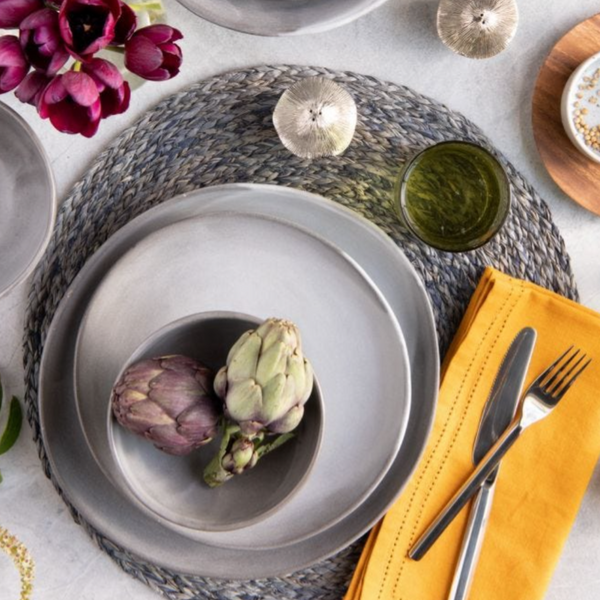 Create a stylish table setting with the Marcus Cement Dinnerware Set of 4 by Blue Pheasant: gray organic plates (microwave safe) topped with artichokes, yellow napkins with cutlery, green sauce in a bowl, purple flowers, and woven placemats on a light surface.