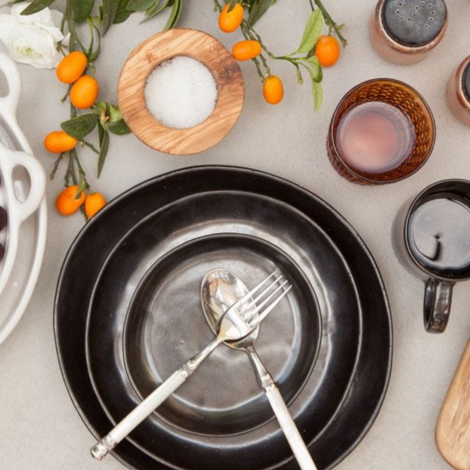 A table setting with Blue Pheasant's Marcus Black Dinnerware Set of 4, featuring black plates with organic shapes, a crossed fork and spoon, a black mug, brown glass, wooden salt bowl, kumquats on a branch, and a partly visible white dish.