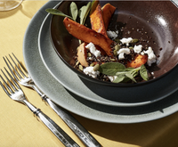 A close-up of roasted sweet potato wedges, quinoa, goat cheese, pumpkin seeds, and sage in a dark brown bowl, set with two forks and textured grey plates from L'Objet’s Terra Seafoam Dinnerware on a yellow tablecloth.