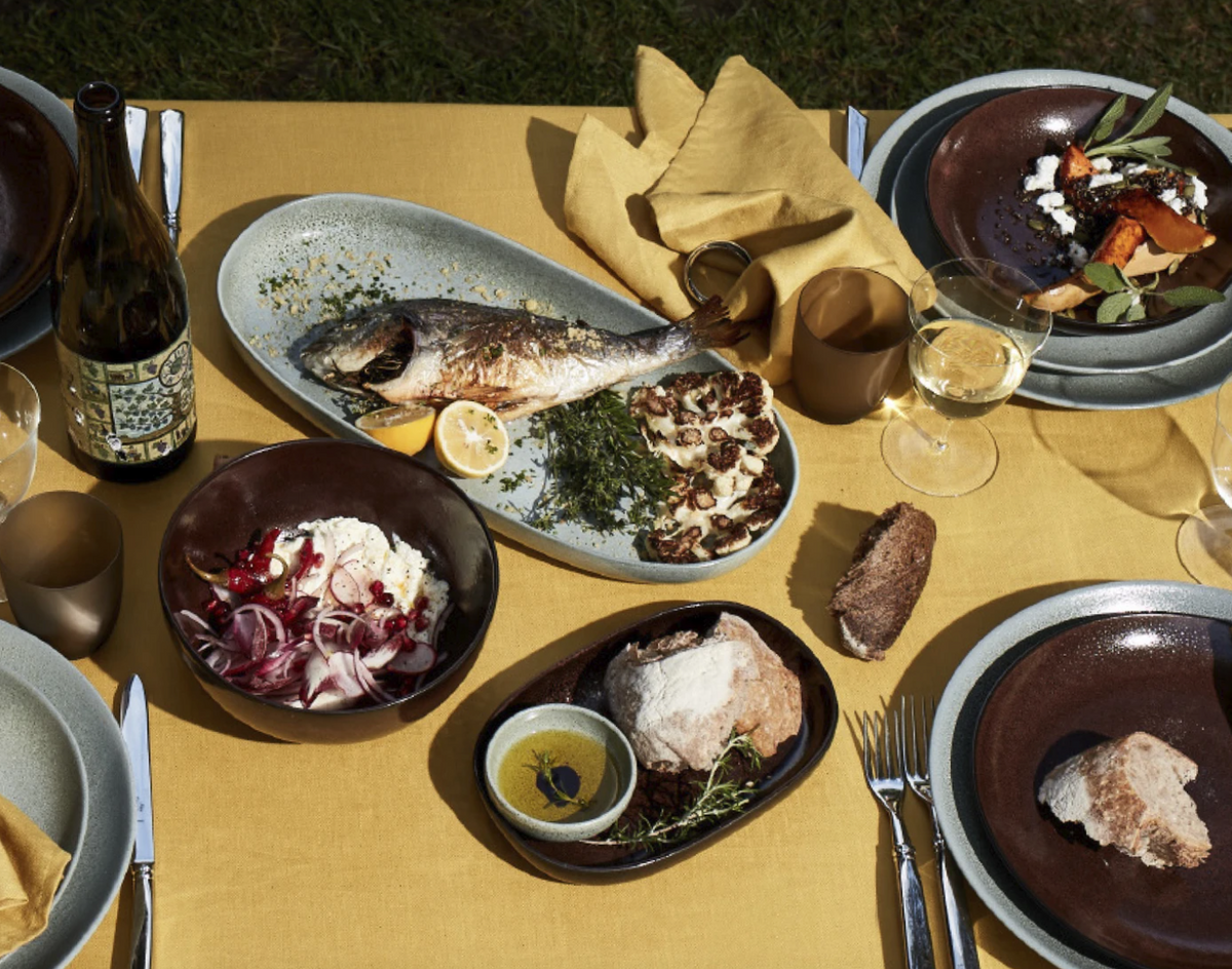 A table set outdoors with a yellow tablecloth features a whole cooked fish, bread, olive oil, salad, roasted vegetables, and elegant brown L’Objet Terra Seafoam Dinnerware alongside a wine bottle and glasses of white wine.