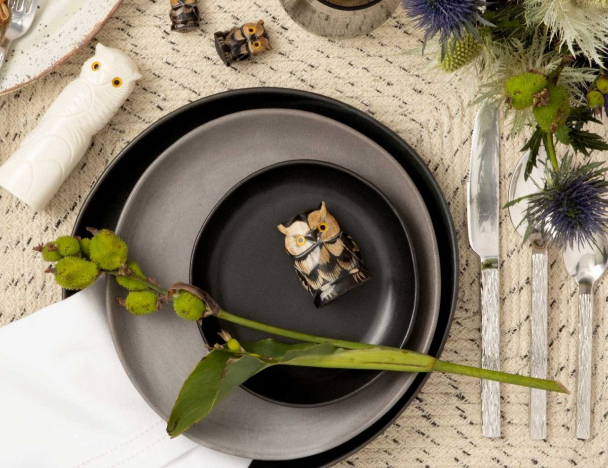 A table set with Blue Pheasant’s Marcus Cement Dinnerware Set of 4 in black and gray, silver cutlery, a small ceramic owl on the top plate, a green plant on a white napkin, decorative greenery, and owl-shaped salt and pepper shakers.