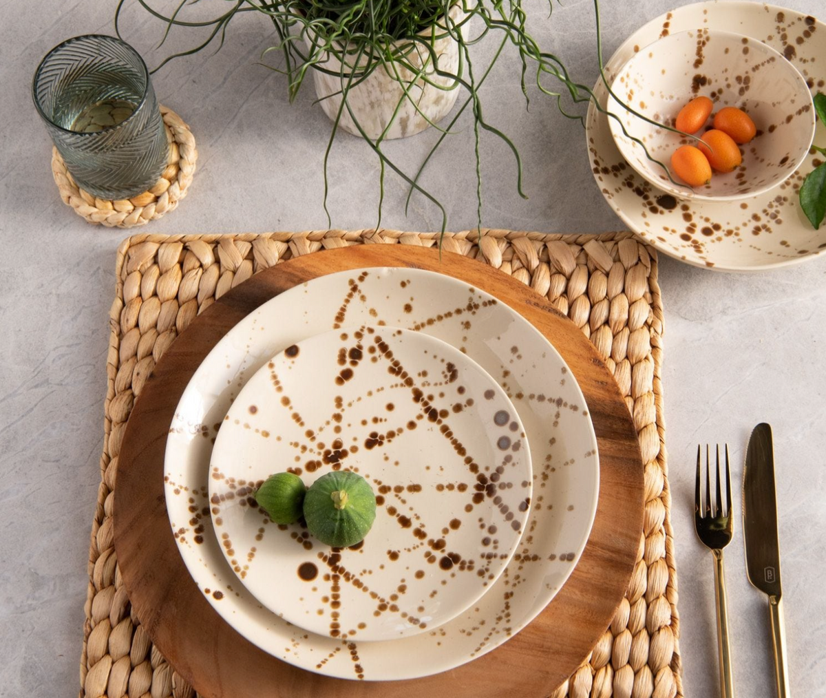 A table setting features Blue Pheasant's Delaney Bronze Splatter Dinnerware—handmade stoneware plates, a matching bowl with oranges, woven placemat, wooden charger, gold utensils, and a potted plant.
