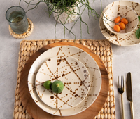 A table setting features Blue Pheasant's Delaney Bronze Splatter Dinnerware—handmade stoneware plates, a matching bowl with oranges, woven placemat, wooden charger, gold utensils, and a potted plant.