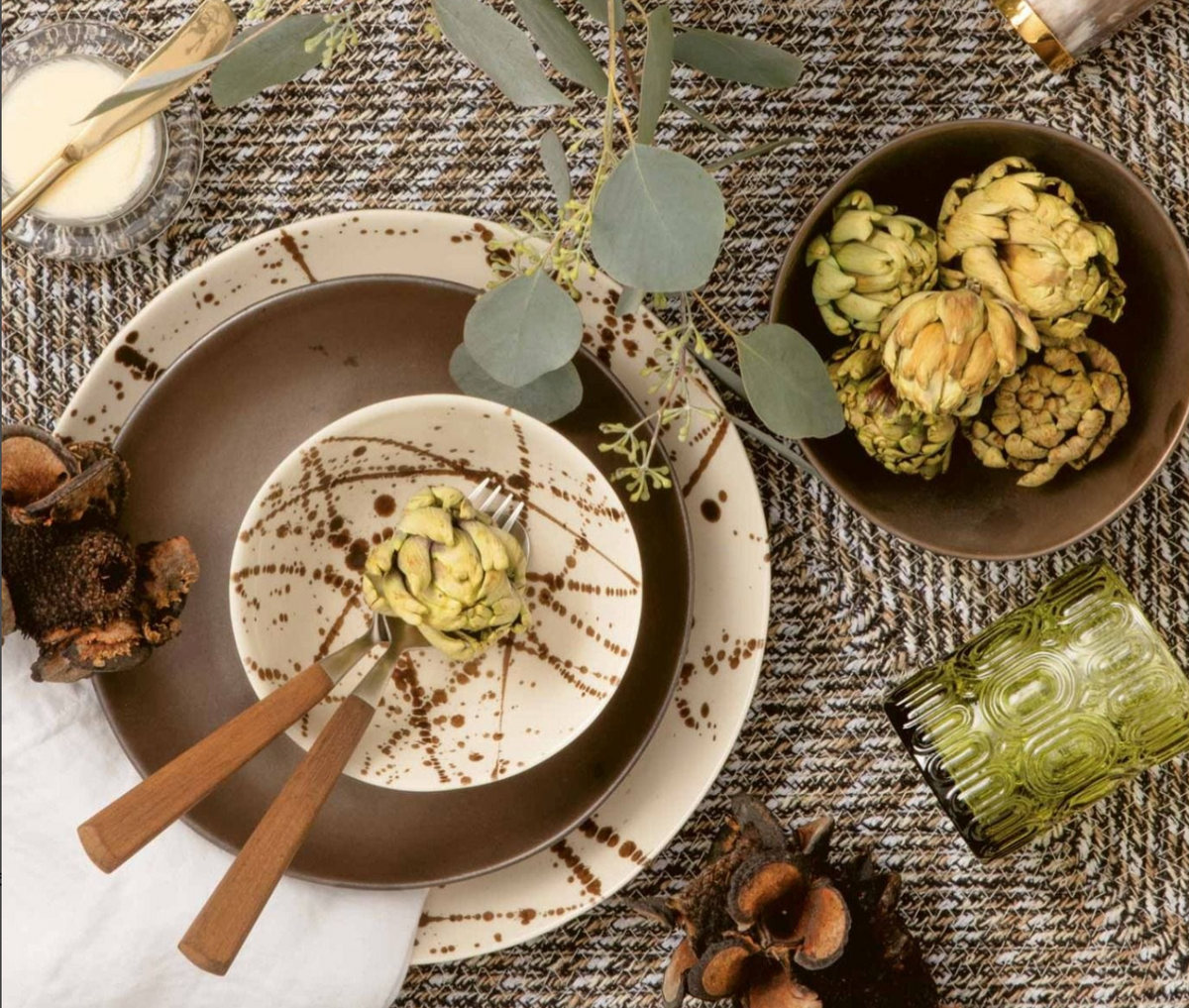 A table setting with Blue Pheasant's Delaney Bronze Splatter Dinnerware, artichokes on plates and bowls, wooden-handled cutlery, a green glass, dried botanicals, and eucalyptus leaves atop a textured cloth.
