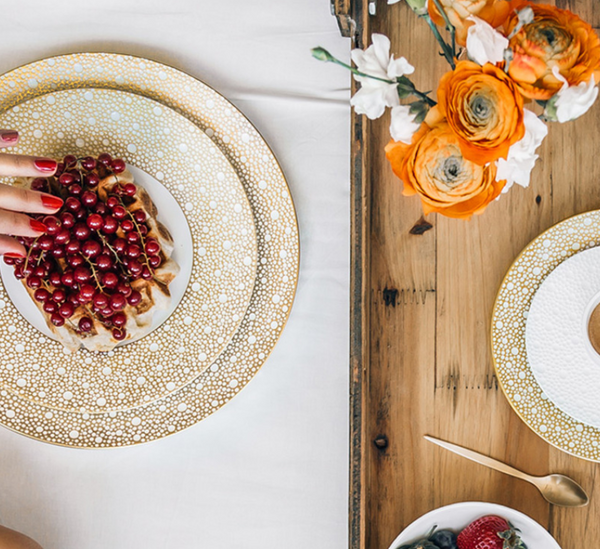 A hand with red nail polish reaches for a waffle topped with red currants on a Bernardaud Ecume Dinnerware Mordore porcelain plate, beside orange flowers and berries on a wooden table.