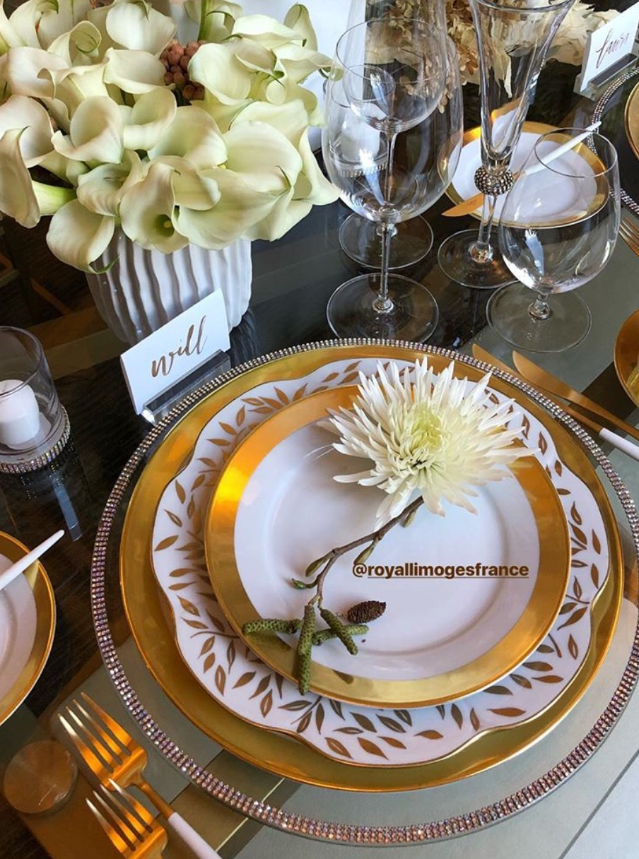 A formal table is set with Danielle Gold Dinnerware by Royal Limoges, elegant silverware, glassware, and a white flower on each French porcelain plate. White calla lilies in a vase and a place card marked Will complete the refined look.
