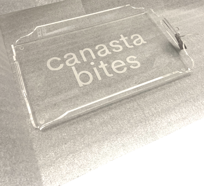 The Eye Candy Deluxe Game Tray Canasta is a clear acrylic serving tray with “canasta bites” in white lowercase letters, displayed on a textured gray surface.