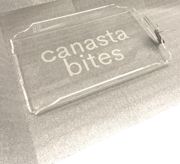 The Eye Candy Deluxe Game Tray Canasta is a clear acrylic serving tray with “canasta bites” in white lowercase letters, displayed on a textured gray surface.